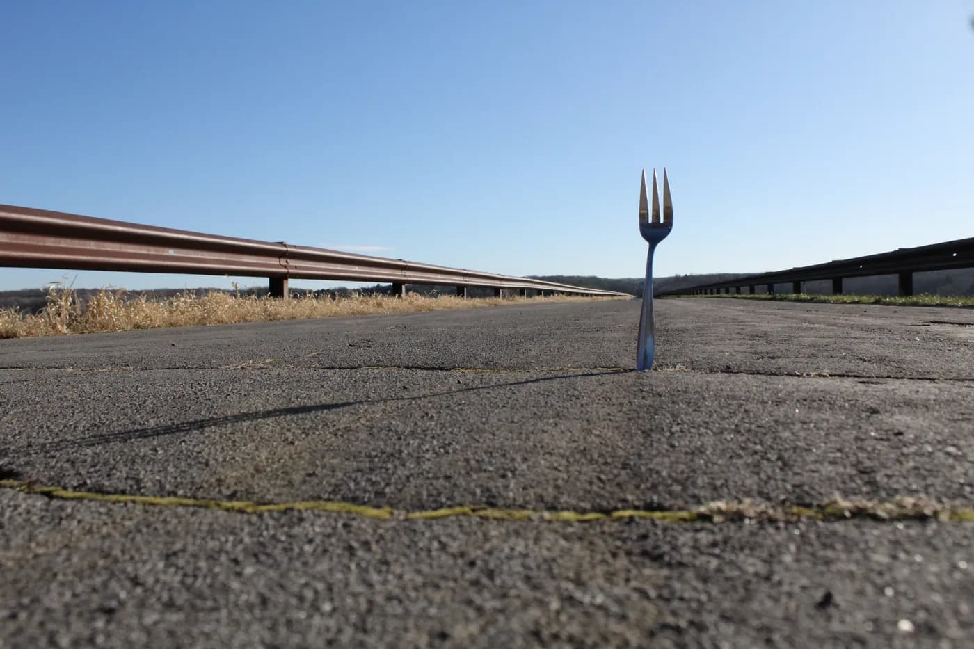 A fork standing upright in the cracked pavement of an empty road, shot from ground level with guardrails leading into the distance