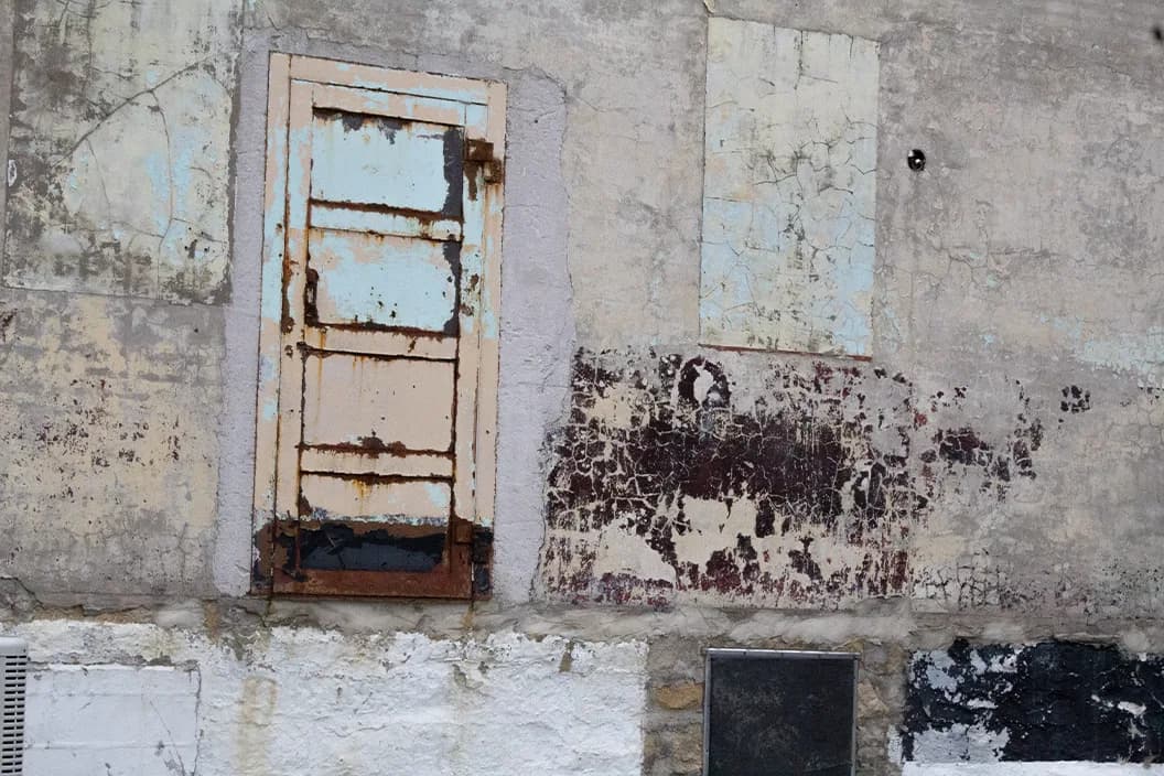 A rusted door set into a flaking, decaying painted wall with tints of light blue